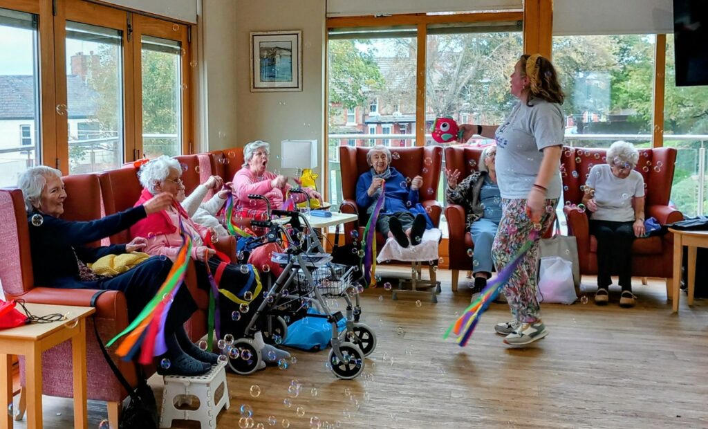 A group of elderly women sit in a lounge waving ribbon wands and blowing bubbles while a woman stands at the front leading the activity.