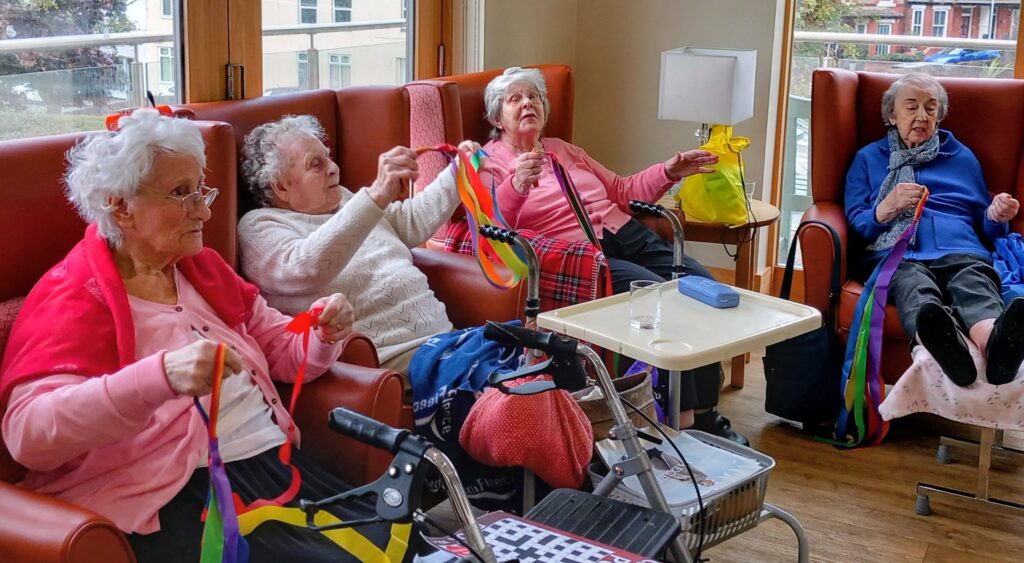 Four elderly women sit in armchairs, holding colourful ribbon streamers, taking part in a group activity in a well-lit room.