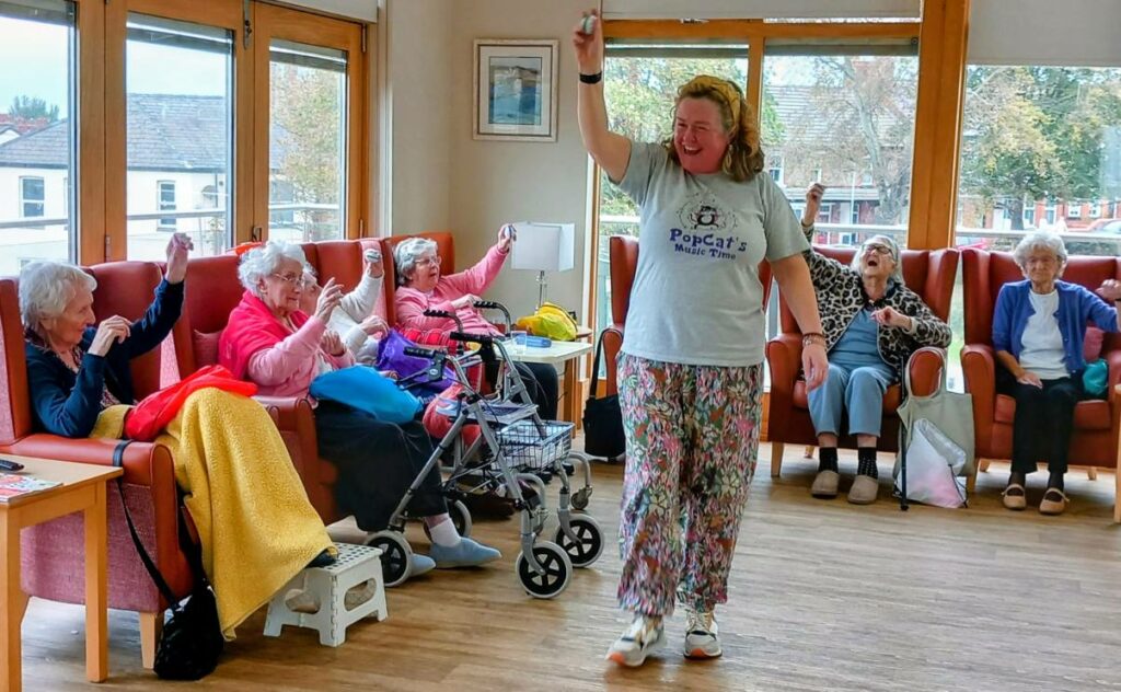 A woman leads a seated group of elderly people in a lively activity, with participants raising their hands and following her movements in a bright, spacious room.