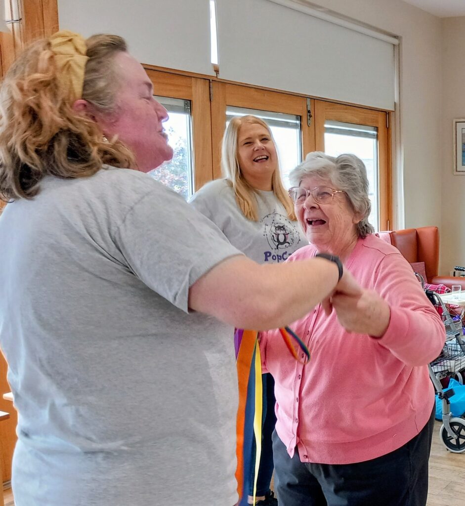 Two women hold hands and seem to dance while smiling, with a third woman standing behind them and watching, all inside a brightly lit room.