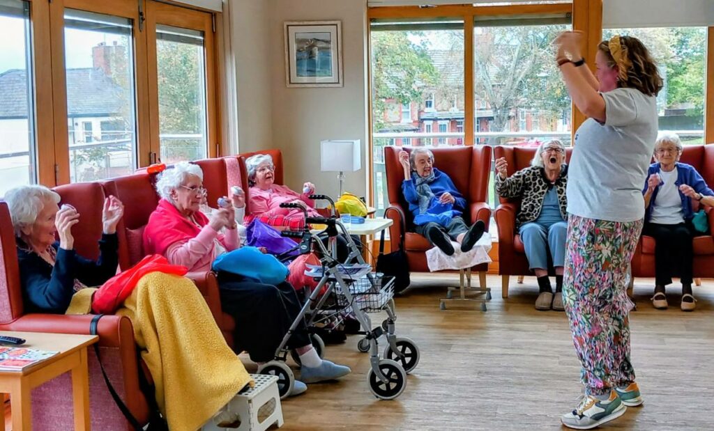 A group of elderly people sit in a common room, taking part in exercise or a group activity led by a woman standing before them with raised hands.