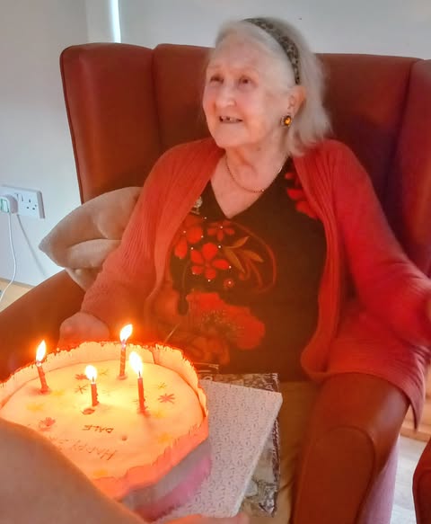 Elderly woman Annie sits in an armchair, smiling as she is presented with a birthday cake topped with four lit candles—Happy Birthday!.