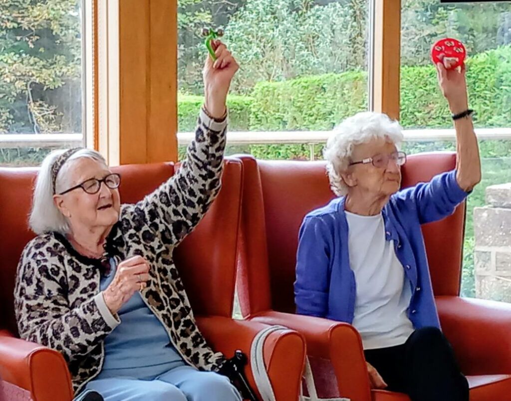 Two elderly women seated in armchairs, each raising a hand and holding a small item—one green, one red—in a bright room with large windows overlooking a garden.