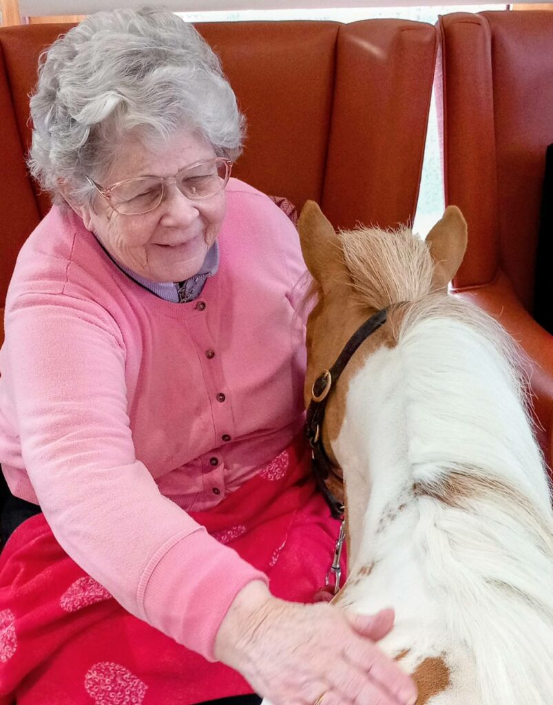 An elderly woman with grey hair and glasses strokes Fudgecake the Pony, a special visitor, while seated on a red cushioned bench during an afternoon event.