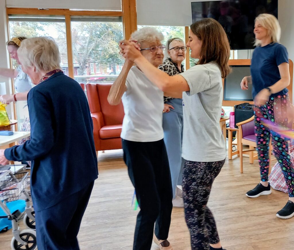 A group of elderly women and a younger woman dance and socialise in a bright, communal room with large windows and comfortable seating.