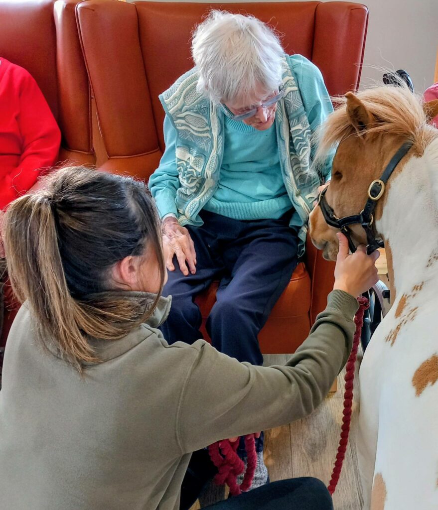 An elderly person sits in a chair, stroking Fudgecake the Pony, a special guest being held by a woman during an afternoon event and therapeutic animal visit.
