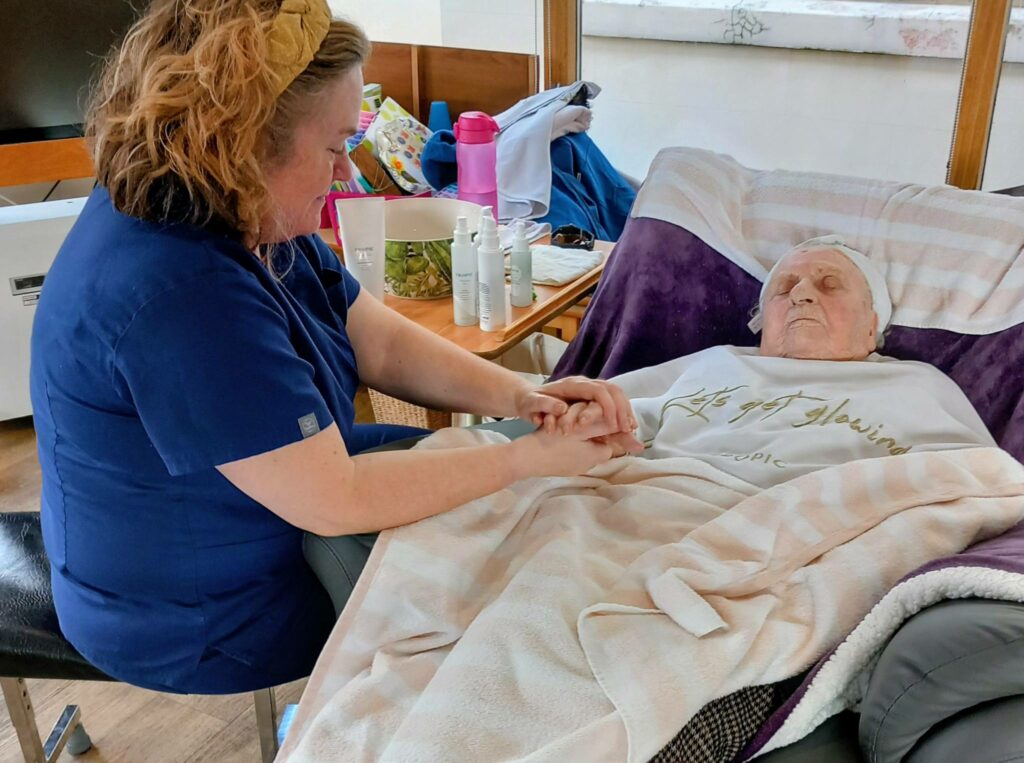 A carer in blue scrubs holds the hand of an elderly person lying in bed beneath a blanket, providing relaxation and care among personal belongings and bottles—a pampering afternoon dedicated to comfort.