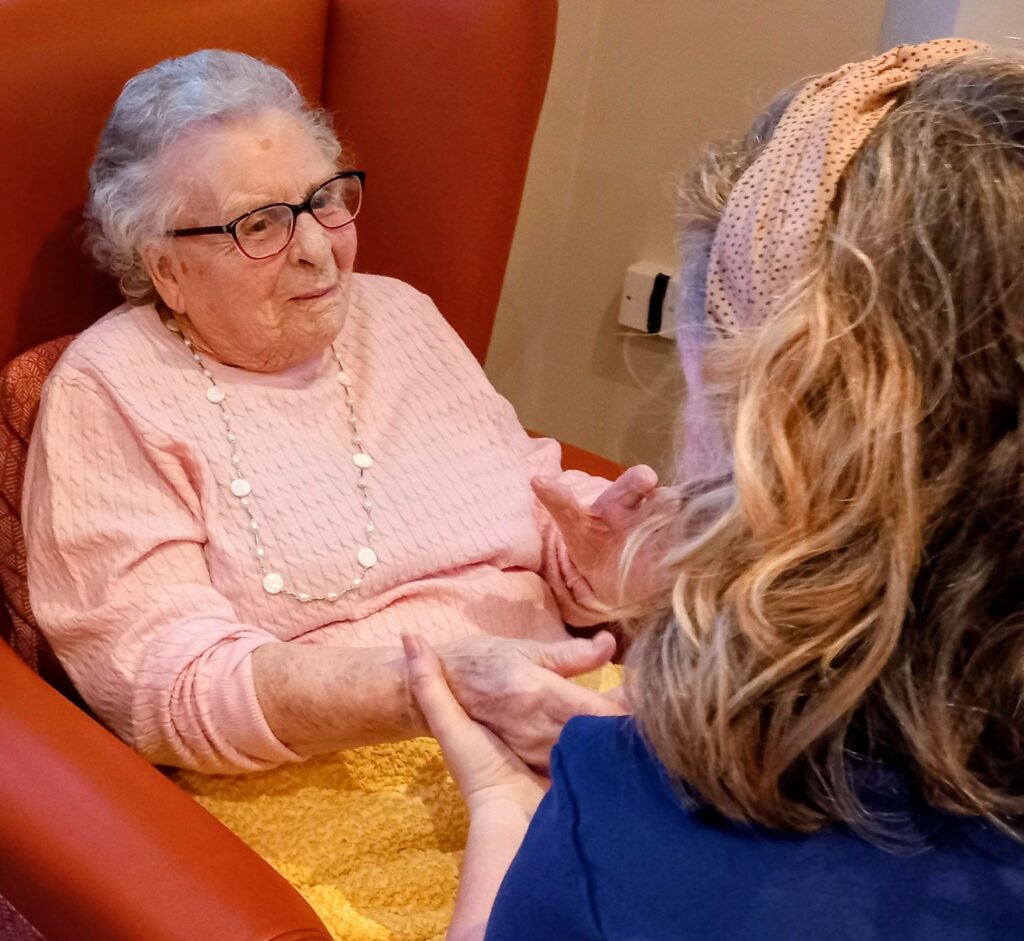 An elderly woman in a pink jumper sits in an orange armchair, enjoying a moment of relaxation as she holds hands and laughs with a younger blonde woman.