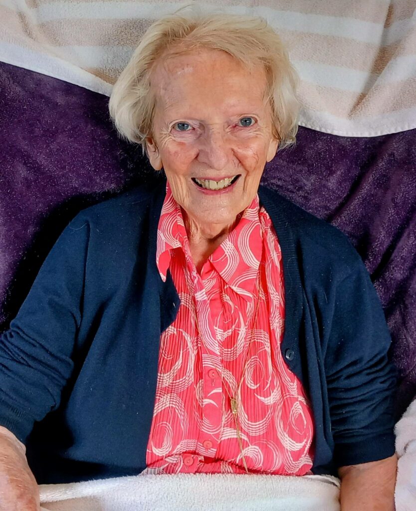 An elderly woman with short white hair smiles at the camera, dressed in a dark cardigan over a bright red patterned blouse. She sits against a purple and striped background, exuding relaxation and contentment.