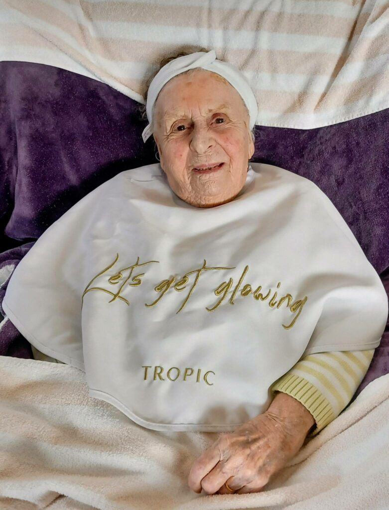 Elderly woman lying on a bed with a headband and a white bib that reads "Lets get glowing TROPIC," smiling at the camera and exuding relaxation and happiness.