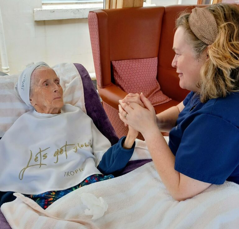 An elderly woman lies in bed wearing a bib that reads Lets get glowing, while a carer sits beside her, holding her hands and smiling, providing an afternoon of relaxation and comfort.
