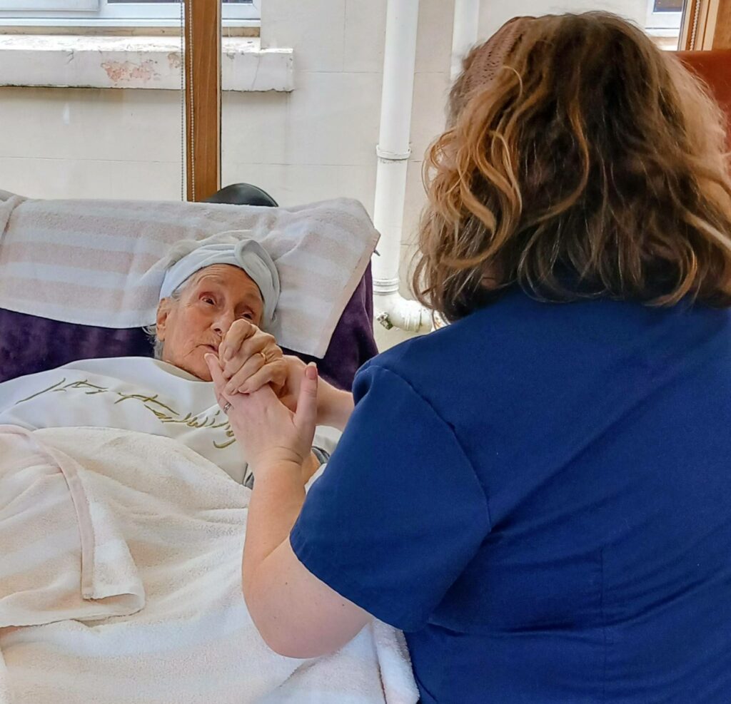 A woman in a nursing uniform holds the hand of an elderly woman, offering relaxation and comfort as she lies in bed, covered with a blanket and a towel.