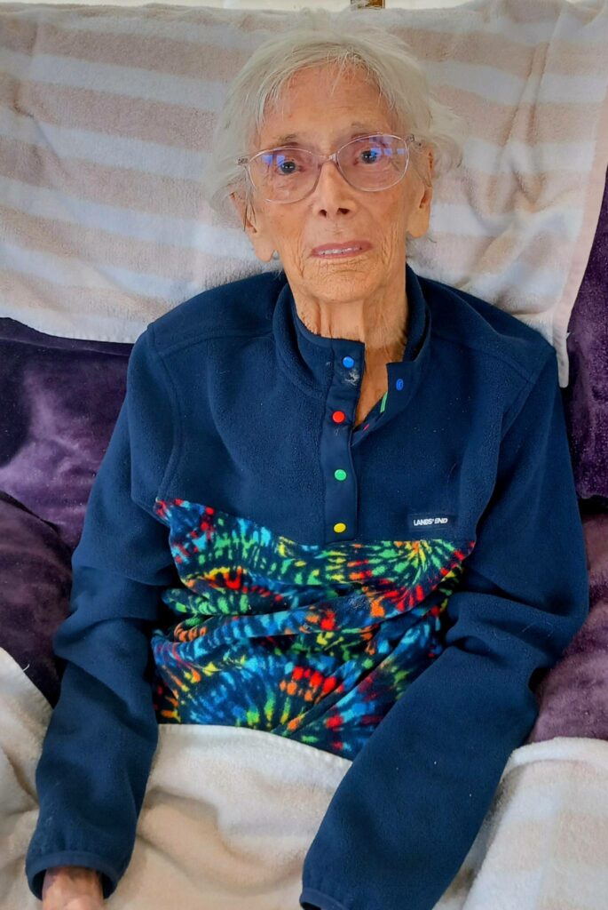 An elderly person with white hair and spectacles sits on a sofa, enjoying relaxation in a colourful tie-dye fleece and covered with a light blanket.