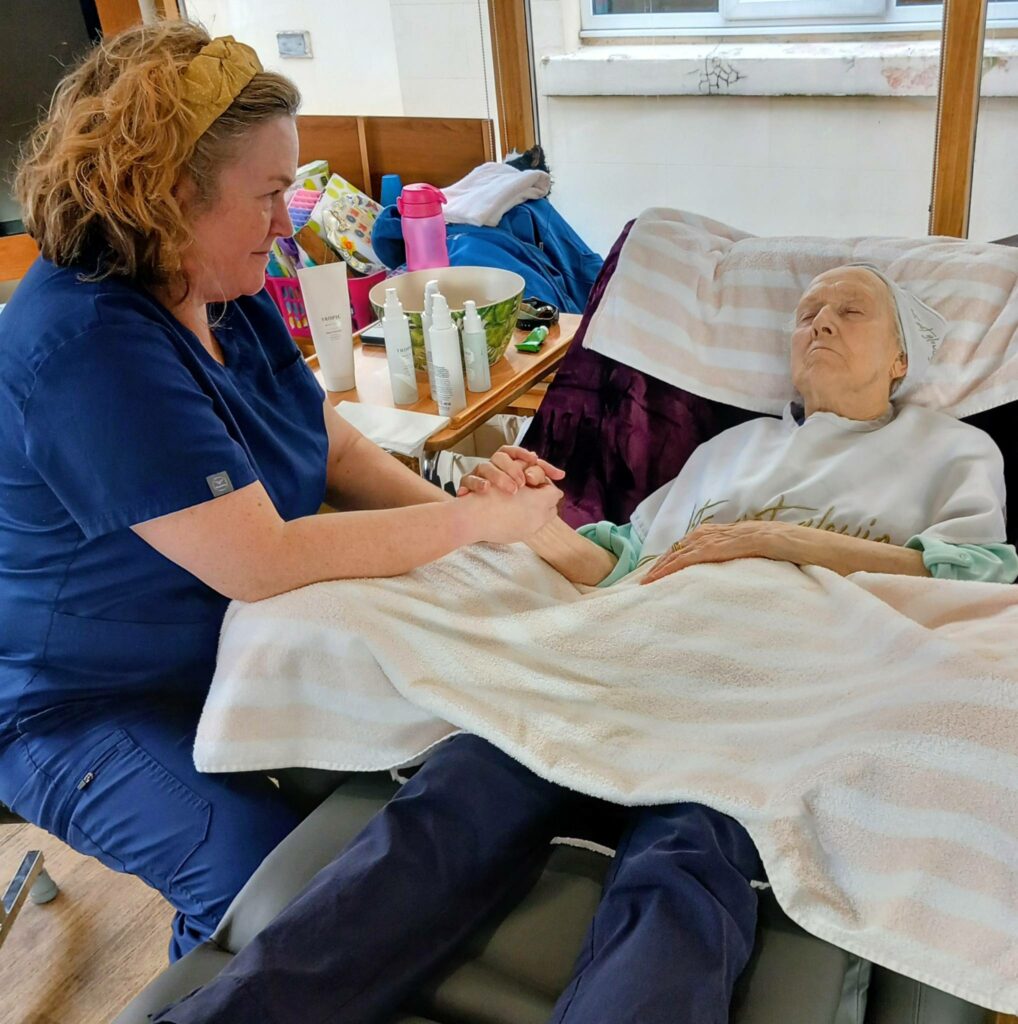 A healthcare worker in scrubs holds hands with an elderly patient lying in a reclined hospital bed, offering relaxation and comfort in the tranquil care home room.