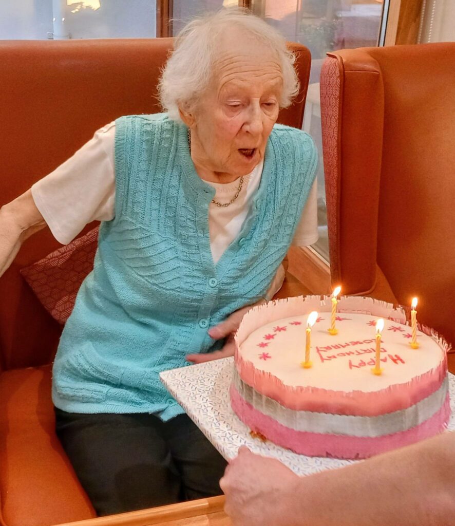 Rosemary, an elderly woman, sits on a bench, blowing out candles on a birthday cake held before her. The sound of Happy Birthday drifts through the air as she makes her wish.