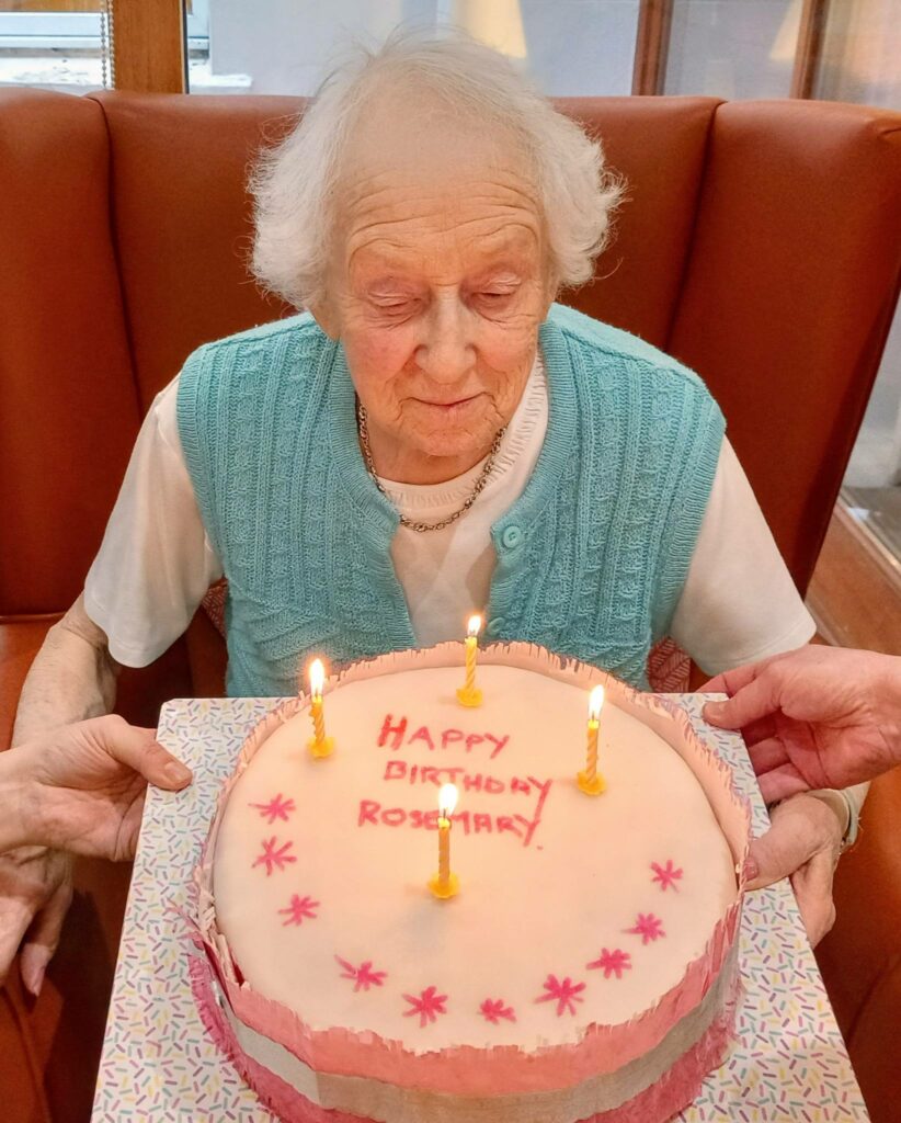 An elderly woman sits in front of a cake with five lit candles and "Happy Birthday Rosemary" written on it, as two hands present the delicious birthday surprise to her.