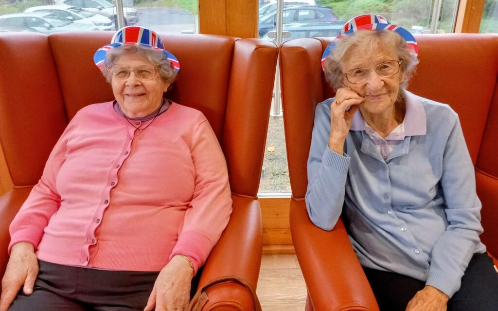 Two elderly ladies wearing red, white and blue hats sit on brown chairs indoors, smiling at the camera and enjoying a spot of British fun.