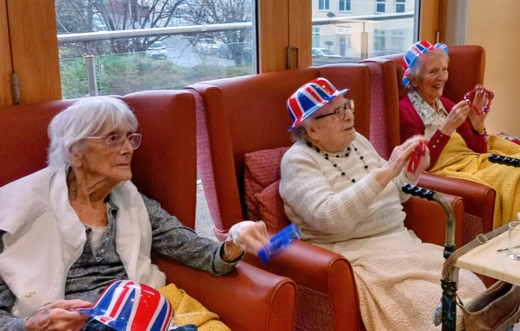 Three elderly women sit in armchairs, sporting red, white, and blue hats, each holding a tambourine and smiling in a brightly lit room, embracing a touch of Boogie Woogie and classic British fun.