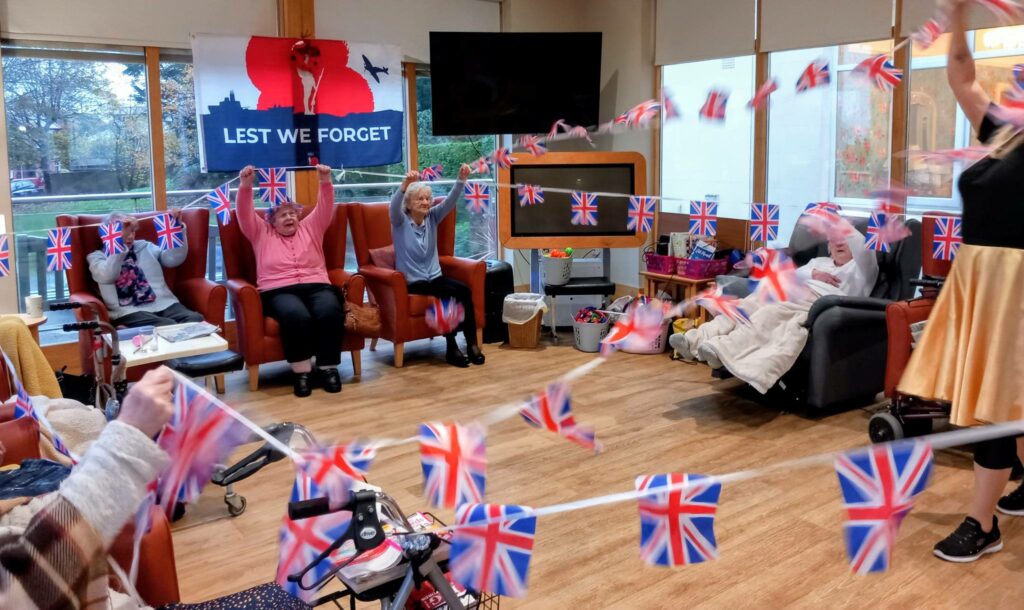 Elderly people seated in a room adorned with Union Jack flags, enjoying British-themed activities; a ‘Lest We Forget’ sign is visible in the background.