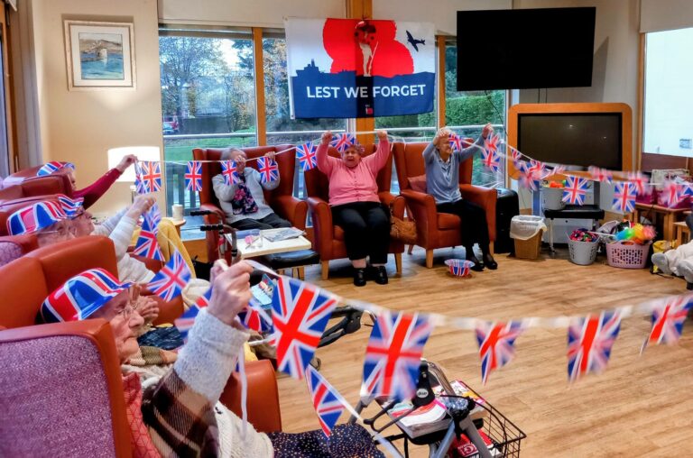 Elderly people seated in a lounge enjoy British festivities, holding Union Jack flags and bunting. A “Lest We Forget” Remembrance banner is displayed on the wall.