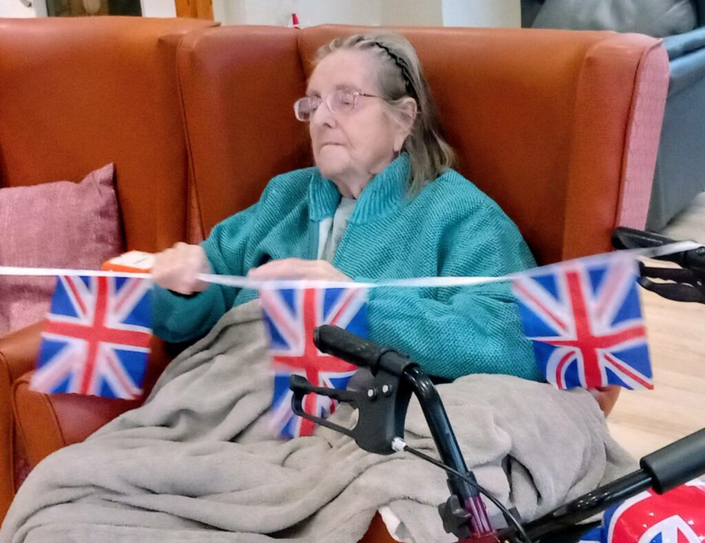 An elderly woman sits in an armchair with a blanket over her legs, holding a string of Union Jack bunting and exuding British fun. A walking frame is visible in the foreground.