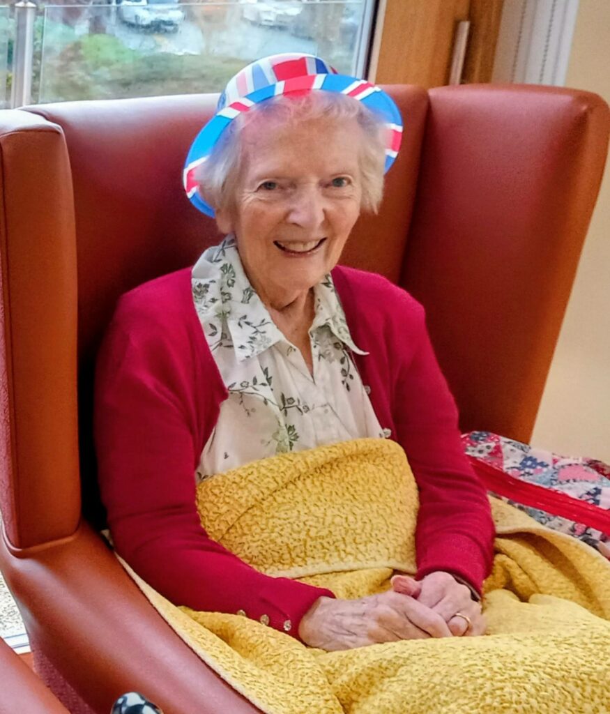 Elderly woman seated in a high-backed chair, sporting a red cardigan and festive hat, with a yellow blanket across her lap, exuding proper British fun.
