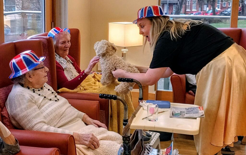 A woman in a festive hat brings British fun as she shows a small dog to two elderly ladies seated in armchairs at a care home. Both women, smiling and wearing matching striped hats, enjoy the lively boogie woogie atmosphere.