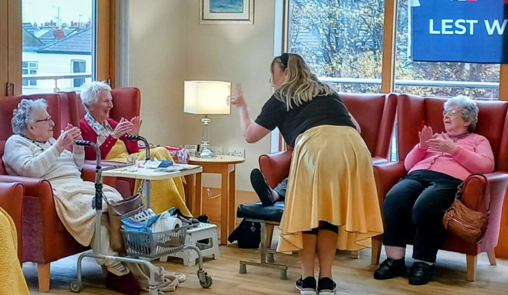 A woman in a yellow skirt adds a dash of British fun as she chats with three elderly ladies in armchairs, one with a walking frame, all gathered in a bright lounge.