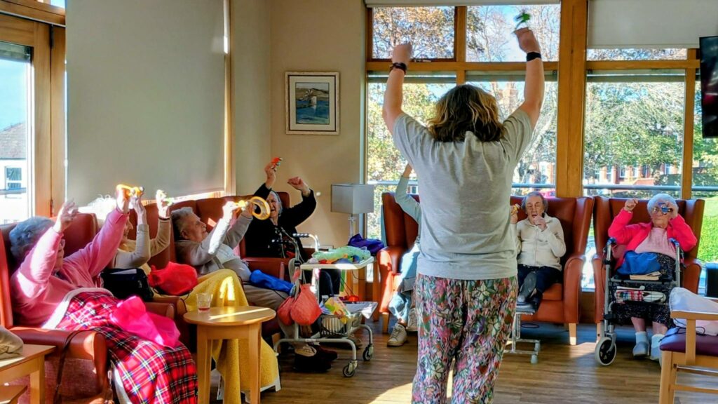 A group of elderly people seated in a sunlit room raise their arms as they follow an instructor leading an exercise session accompanied by lively music.
