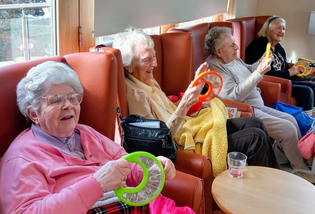 Four elderly women sit in armchairs, smiling and holding colourful plastic rings. Laughter fills the room as they enjoy each other's company. A glass of water and a folded blanket are on the table in front of them.