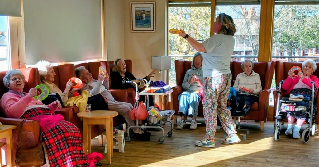 A group of elderly people sit in armchairs, laughing together as they join in an exercise activity led by a facilitator holding a ring, with gentle music playing in the brightly lit room.