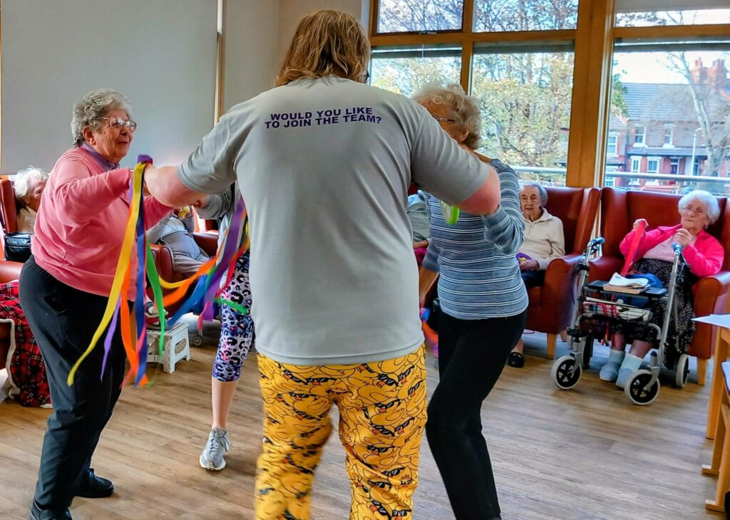 A group of pensioners and a staff member dance in a circle with colourful ribbons to lively music, while other pensioners watch from chairs in the background of a bright room.