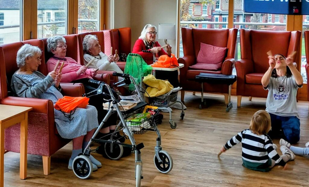 Several elderly women sit in armchairs, while a woman and child sit on the floor before them, sharing laughter and music together in a bright communal room.