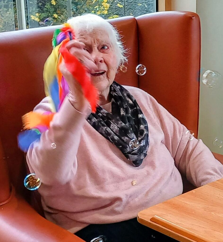 Elderly woman seated in a chair smiles as she waves a colourful ribbon toy, with bubbles floating around her and cheerful music playing in the background.