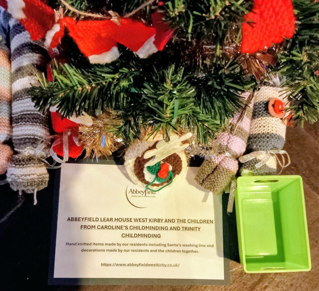 Knitted Christmas decorations hang on a tree at St Hildeburgh’s Festival above a sign explaining they were made by Abbeyfield Lear House residents and local childminding groups, with a small green tray nearby.