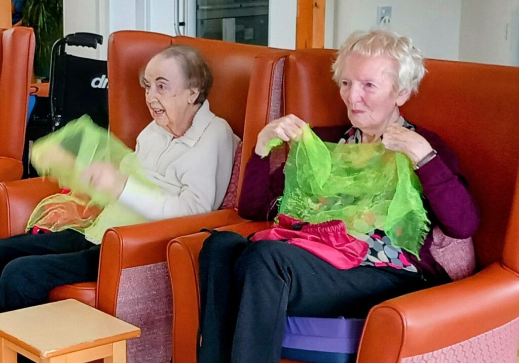 Two elderly women sit in armchairs, each holding a sheer green scarf, laughing together as they take part in a cheerful indoor activity.