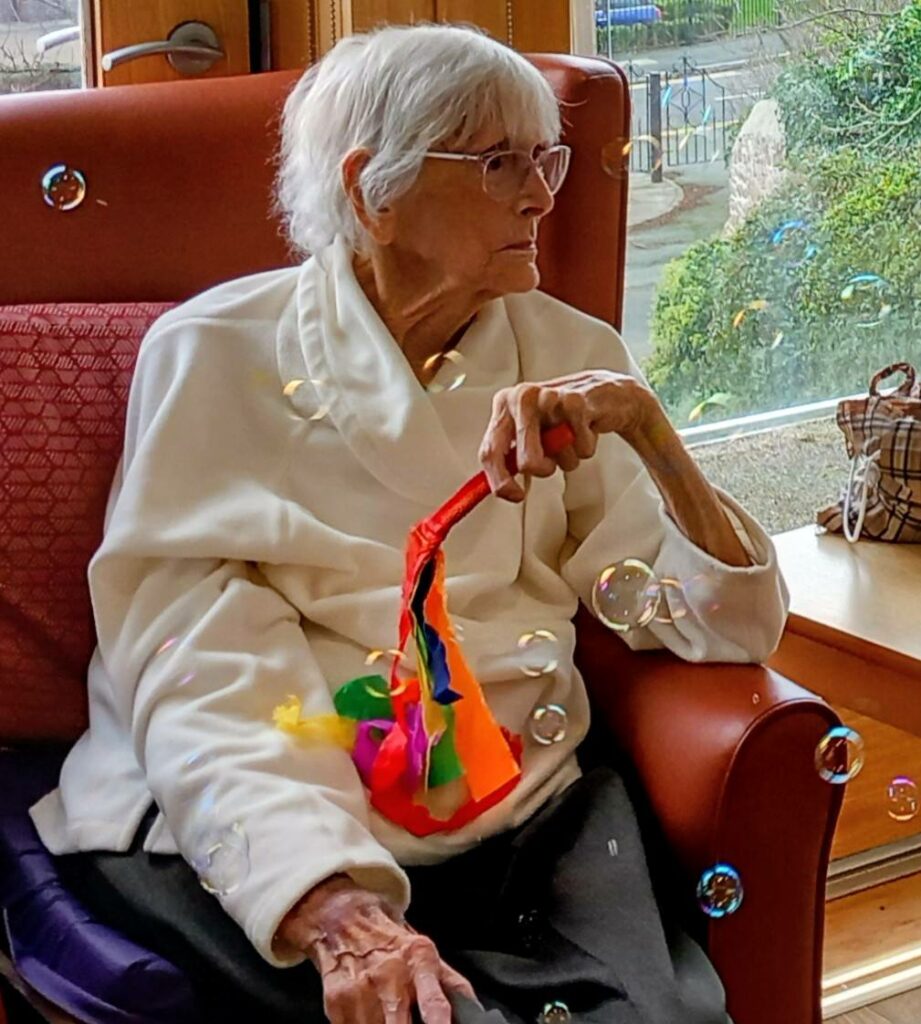 Elderly woman with short white hair and glasses sits in an armchair by a window, holding a colourful ribbon wand, as bubbles float around her to the gentle sound of music.