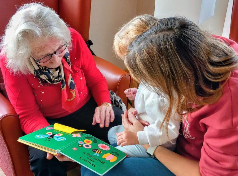 An elderly woman reads a colourful children’s book to a young child sitting on a woman’s lap, all in red chairs, their laughter echoing like joyful music throughout the room.