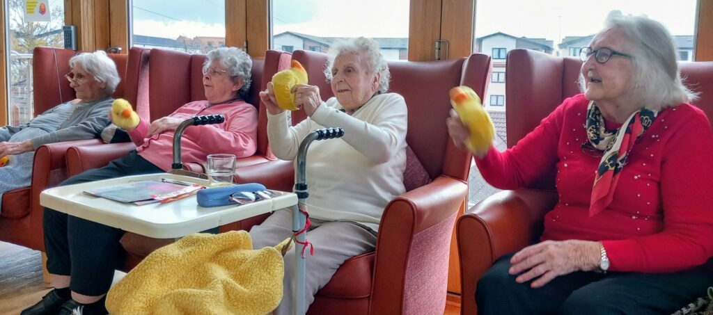 Four elderly women seated in armchairs take part in an exercise activity, each holding a yellow plush dumbbell-shaped object while gentle music plays softly in the background.