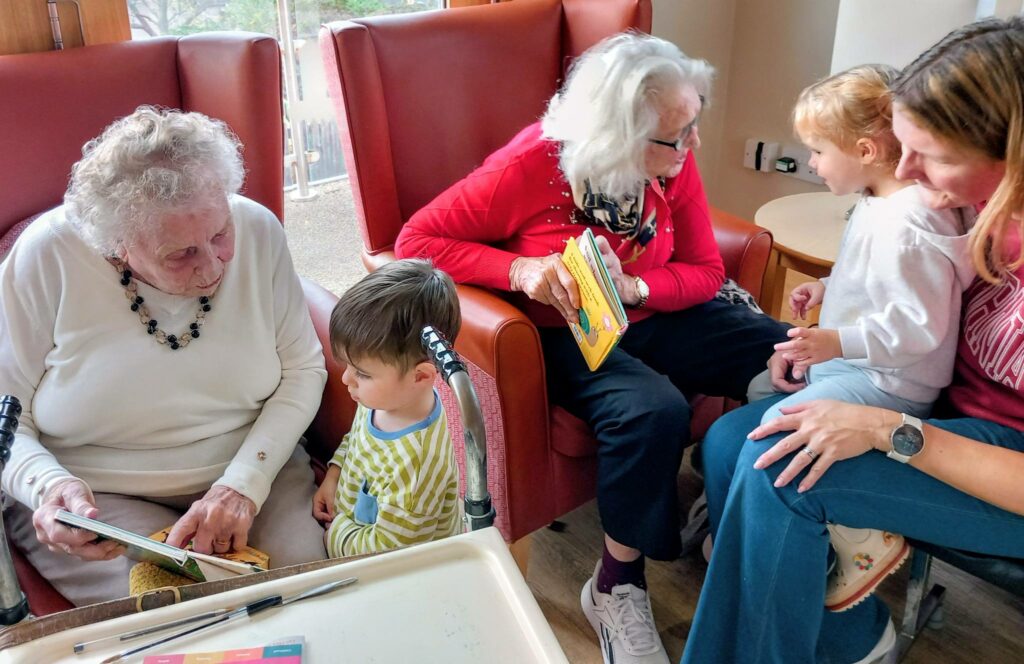 Two elderly women sit in armchairs reading books to two young children, while an adult woman sits close by. Laughter fills the room as the group enjoys a shared reading session indoors.