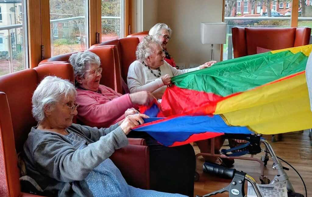 Four elderly women seated in armchairs indoors share a laugh as they play with a colourful parachute, their Joyful Popcats spirit shining through.