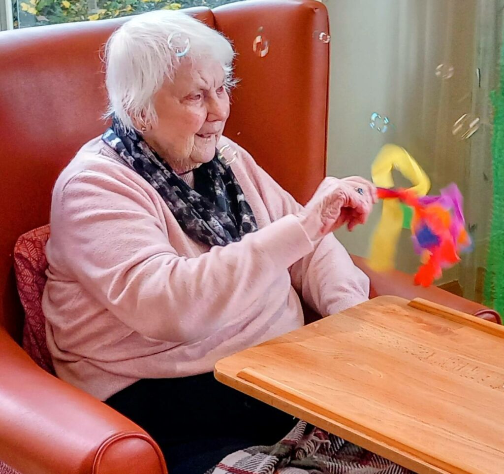 Elderly woman seated in a chair waves a colourful ribbon wand, bubbles floating around her and a wooden tray in front, as Joyful Popcats music fills the air.