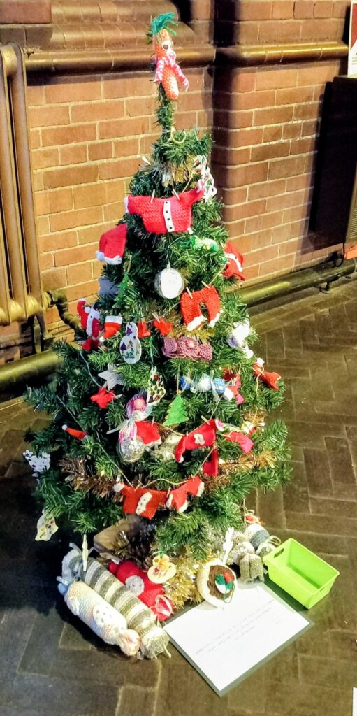 A carefully decorated Christmas tree adorned with handmade knitted ornaments, with stuffed animals at its base, stands on a brick-tiled floor at St Hildeburgh’s Festival, accompanied by a sign and green container.
