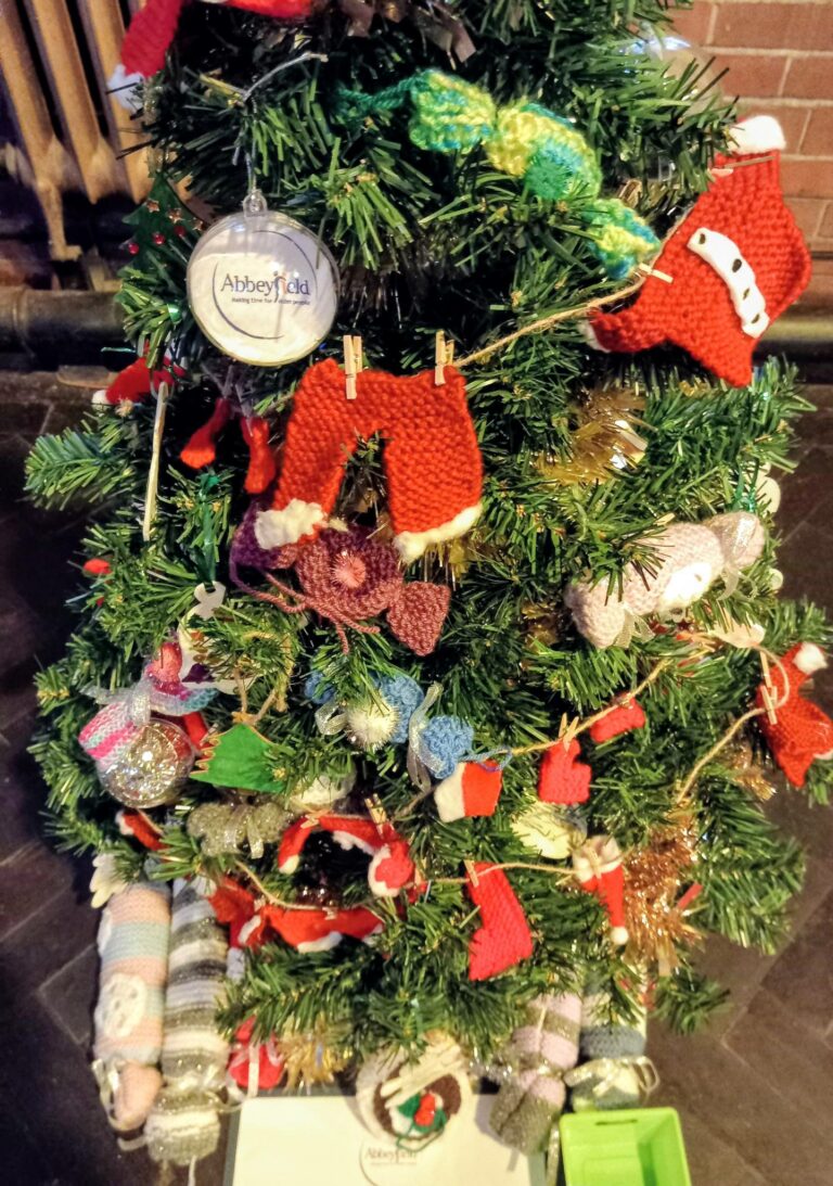 A Christmas tree at the St Hildeburgh’s Festival is adorned with a variety of knitted decorations, including garments, stockings, animals, and a round Abbeyfield bauble—all lovingly handcrafted.