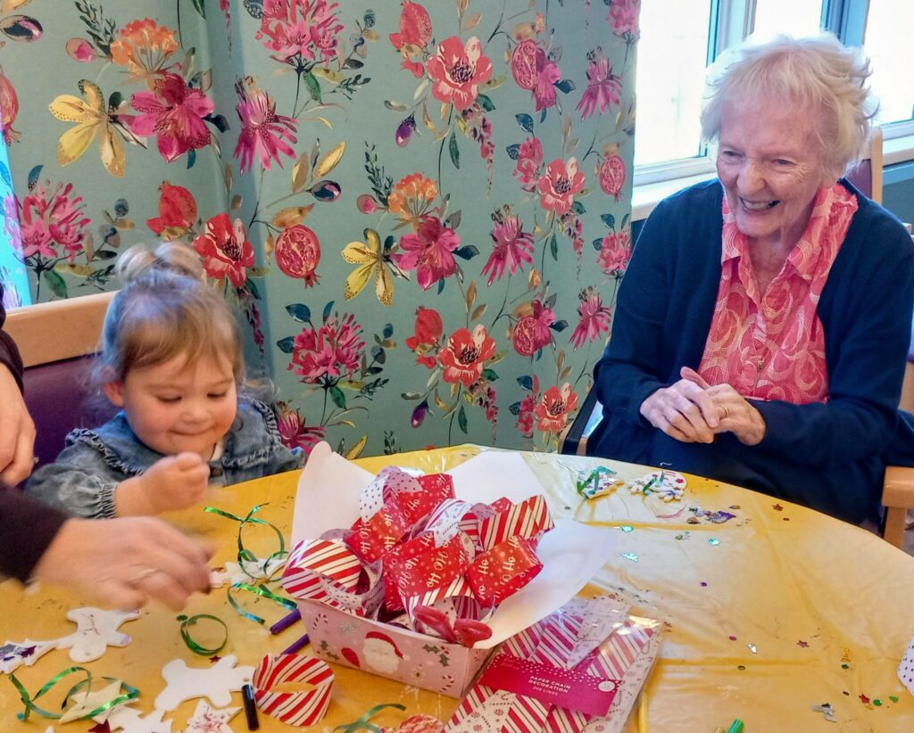 An elderly woman and a young girl sit at a table adorned with gifts and crafts, smiling as they spend time together beneath the Tree Made with Care at St Hildeburgh’s Festival in a brightly decorated room.