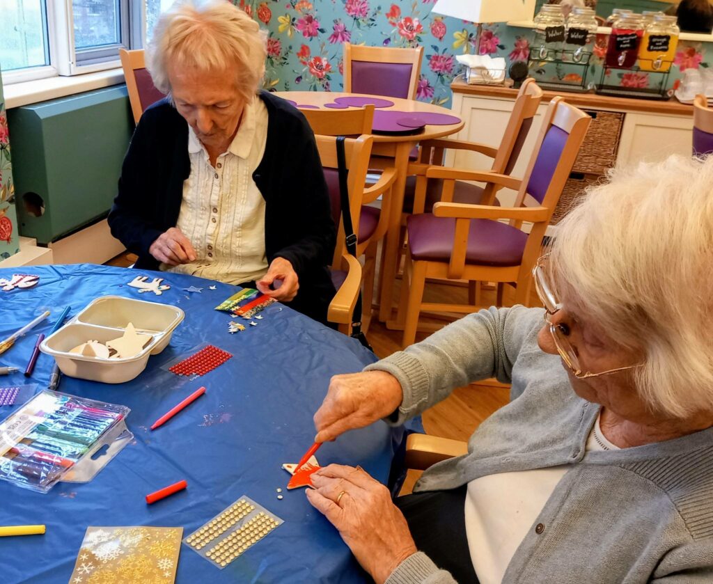 Two elderly women are seated at a table covered with craft materials, engaged in art projects in a brightly decorated room.