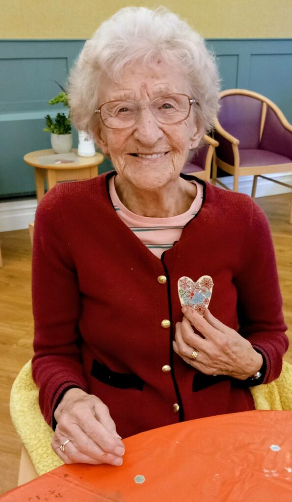 An elderly woman with white hair and glasses smiles as she holds a carefully crafted fabric heart. She sits at a table draped in an orange cloth at St Hildeburgh’s Festival, in a room with green walls and wooden chairs.