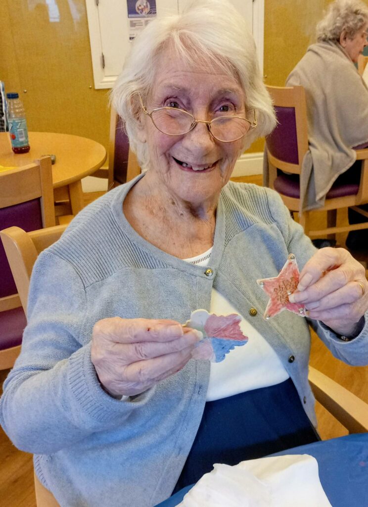 Elderly woman seated at a table, smiling and holding up two decorated paper cut-outs during St Hildeburgh’s Festival, with other people and a festive atmosphere in the background.
