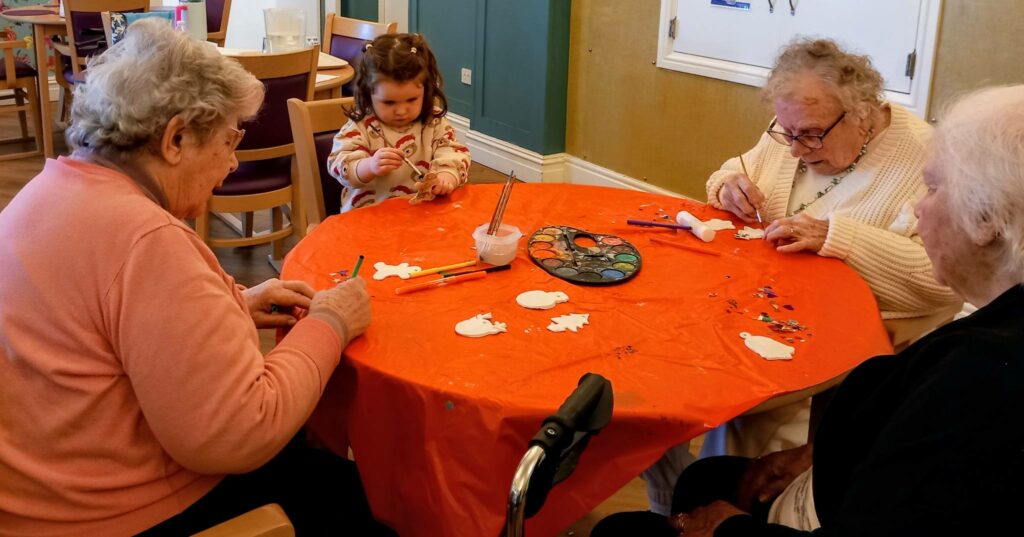 Three elderly women and a young girl sit round a table, painting craft shapes with brushes at St Hildeburgh’s Festival, making decorations for the Tree Made with Care.
