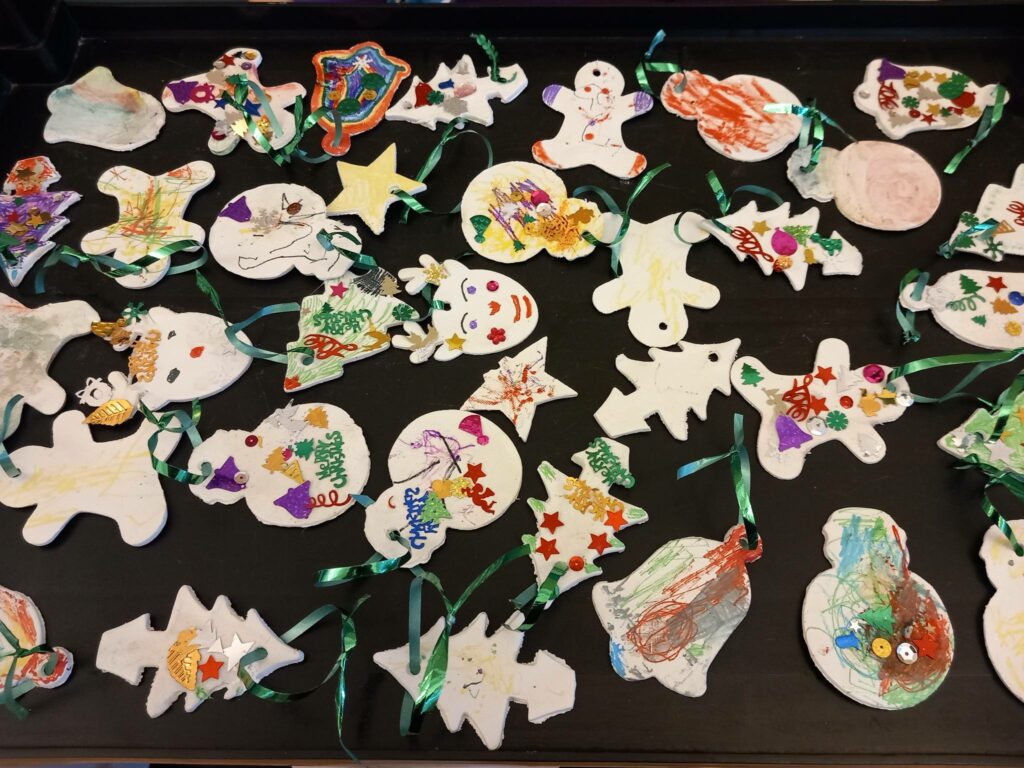 A selection of handmade Christmas decorations, ideal for your tree, adorned with paint, glitter, stickers and ribbons, are displayed on a black surface at St Hildeburgh’s Festival.